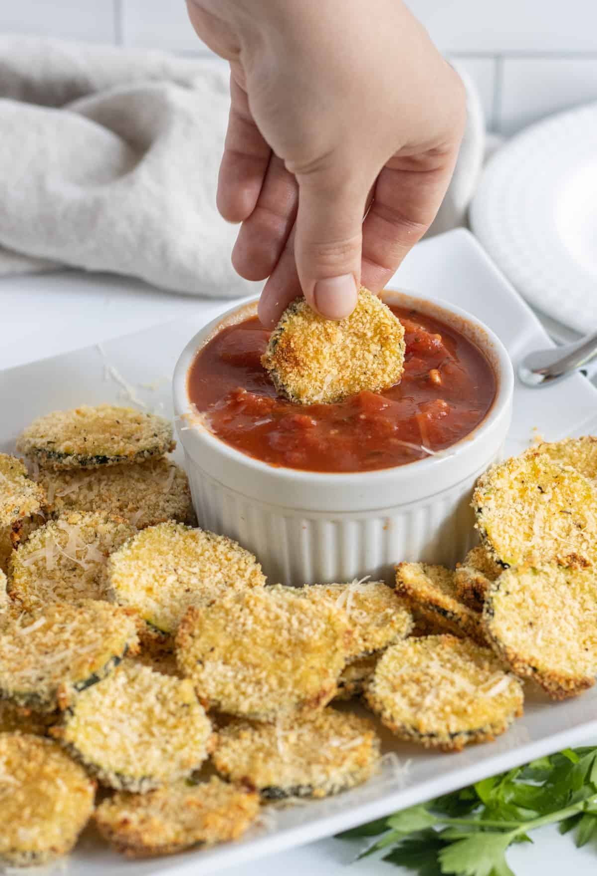 A hand dipping an oven fried zucchini chip into a bowl of marinara.