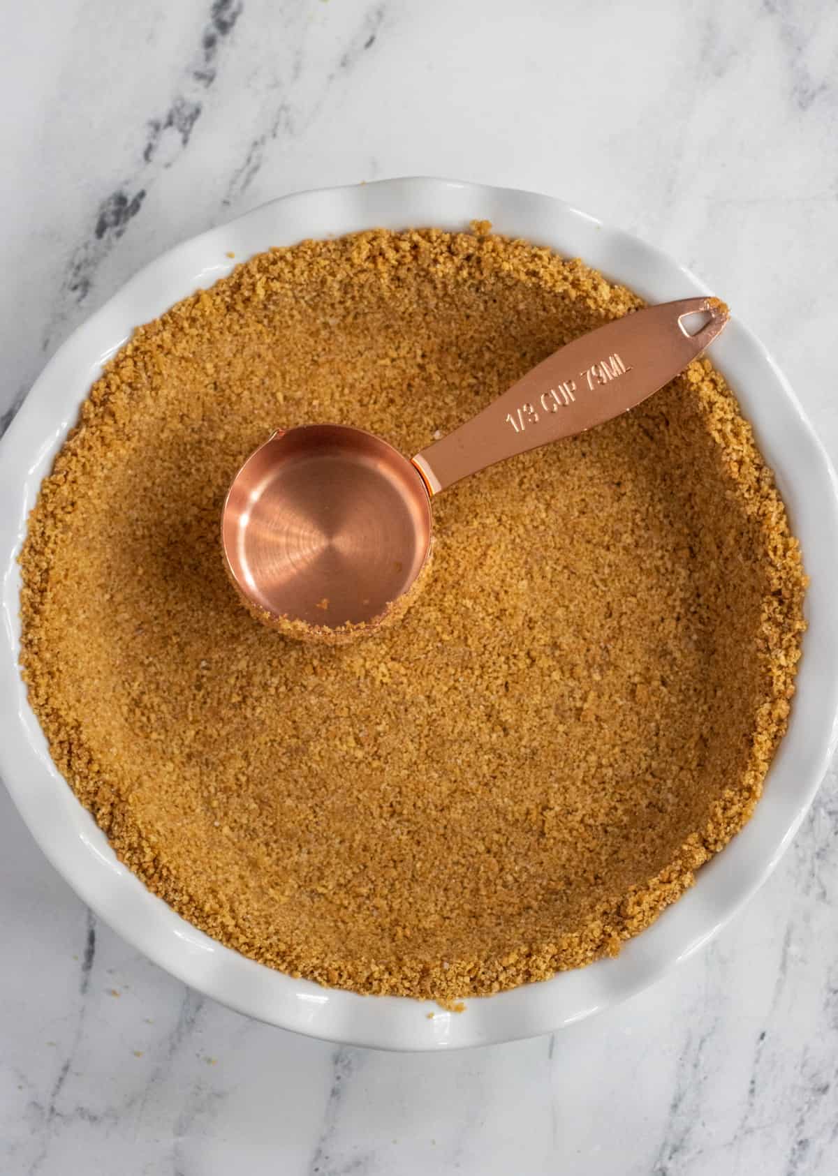 Graham cracker crust being pressed into a pie plate with a measuring cup.