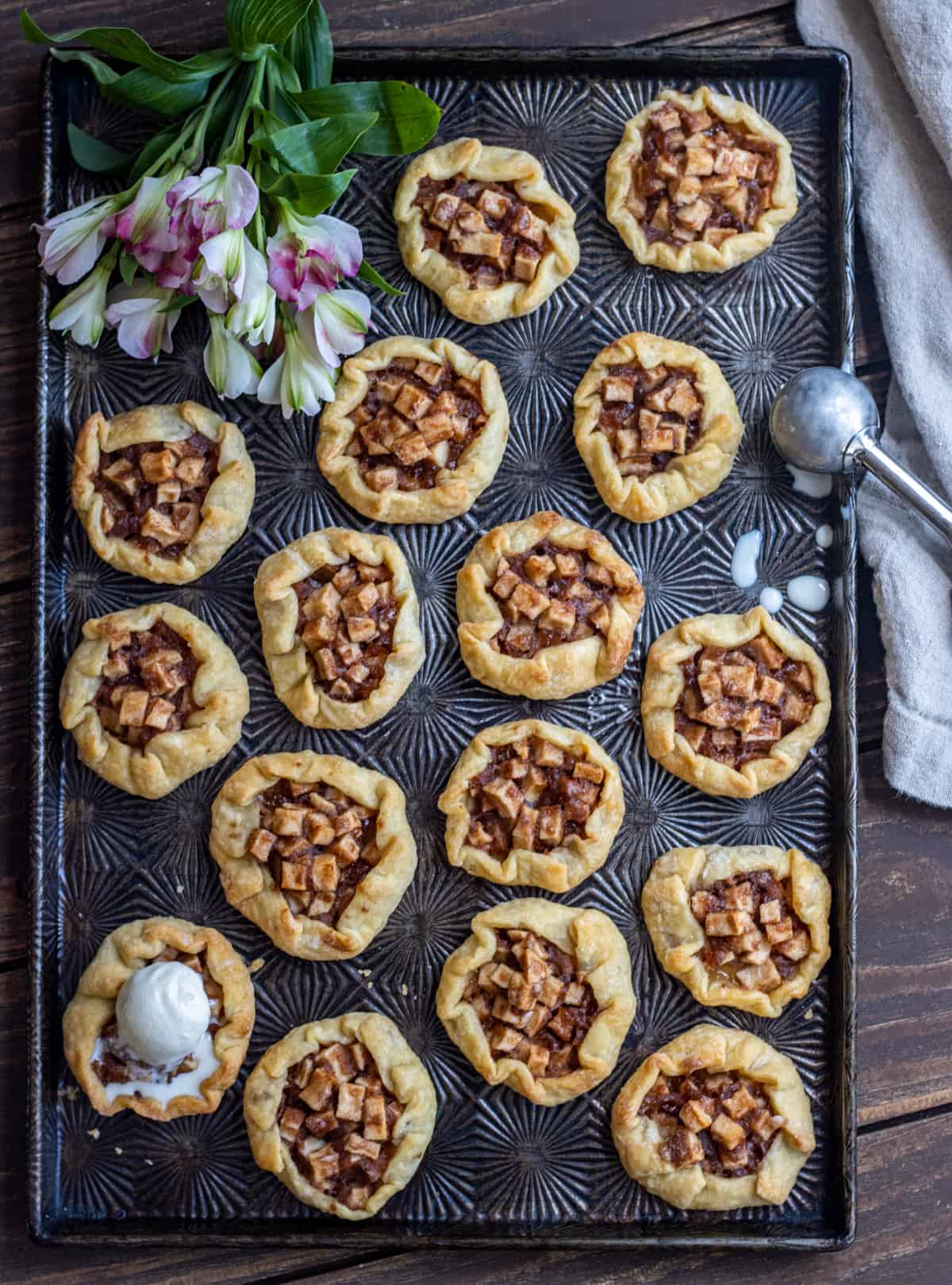 Mini apple galettes on a dark tray with a scoop of ice cream on one galette and flowers in the corner.