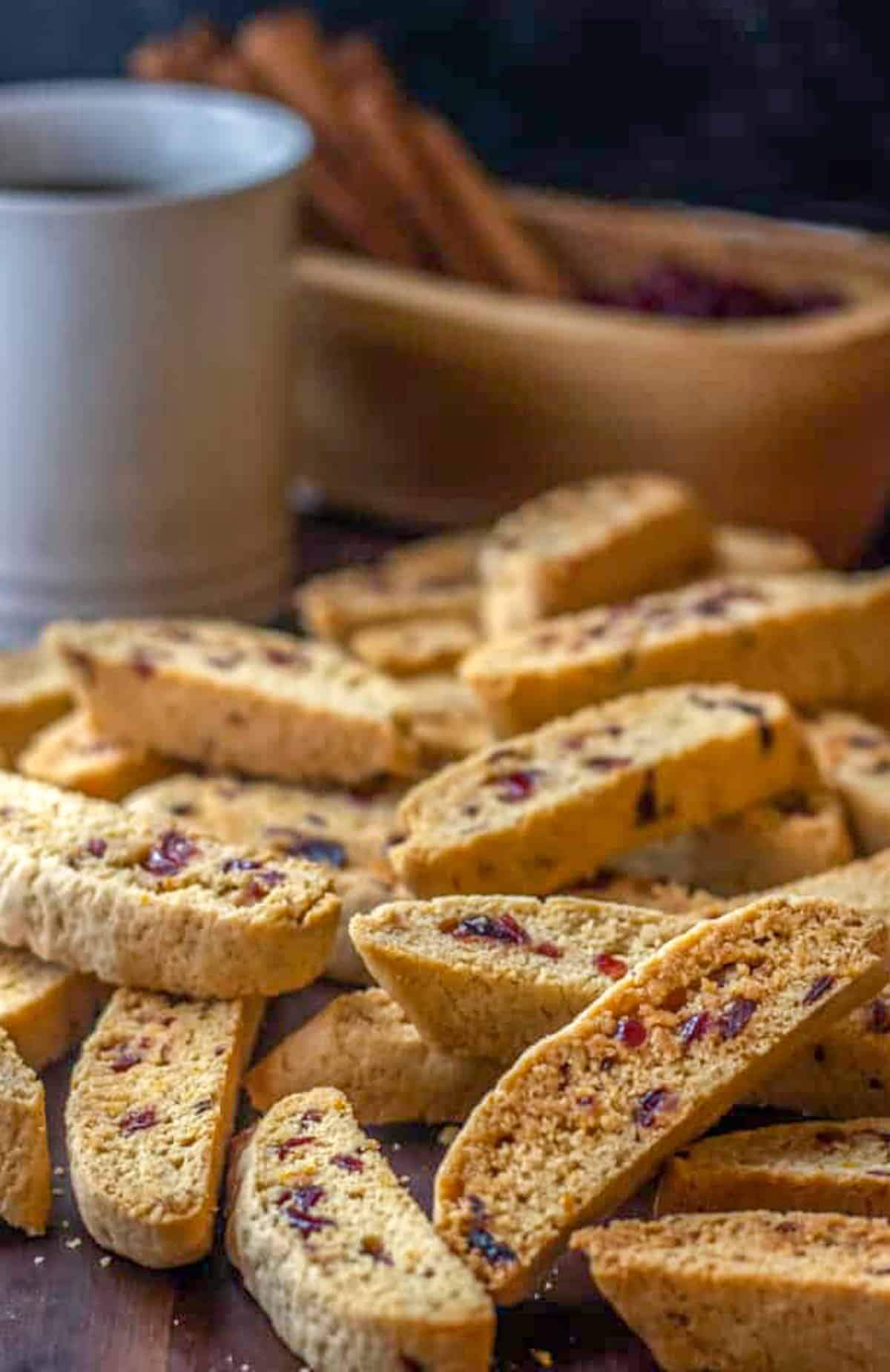 Cranberry orange biscotti piled up in front of a cup of coffee and container of cinnamon sticks.
