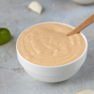 Sriracha aioli in a bowl with lime wedges and garlic cloves all around.