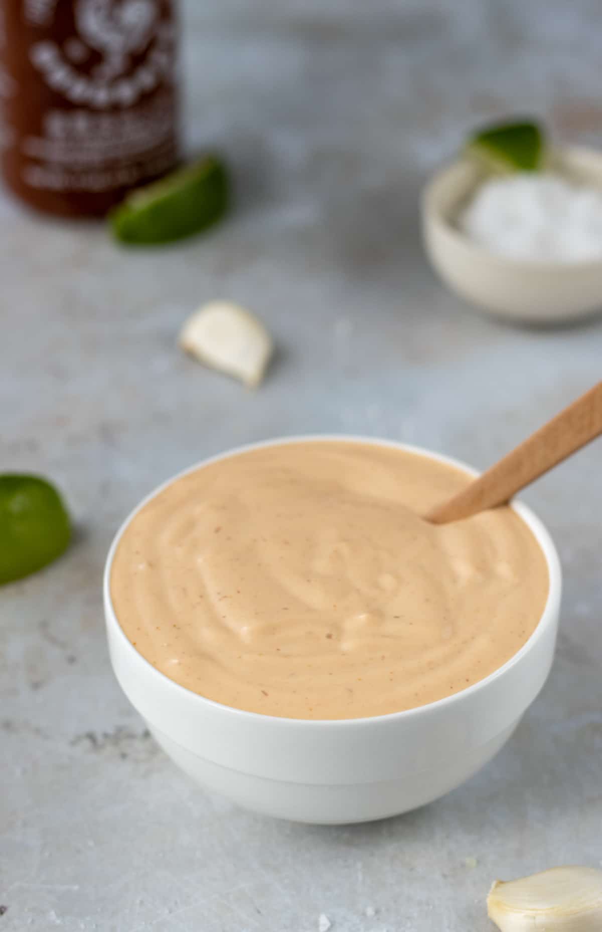 Sriracha aioli in a bowl with lime wedges, garlic cloves, a bowl of salt, and a sriracha bottle in the background.