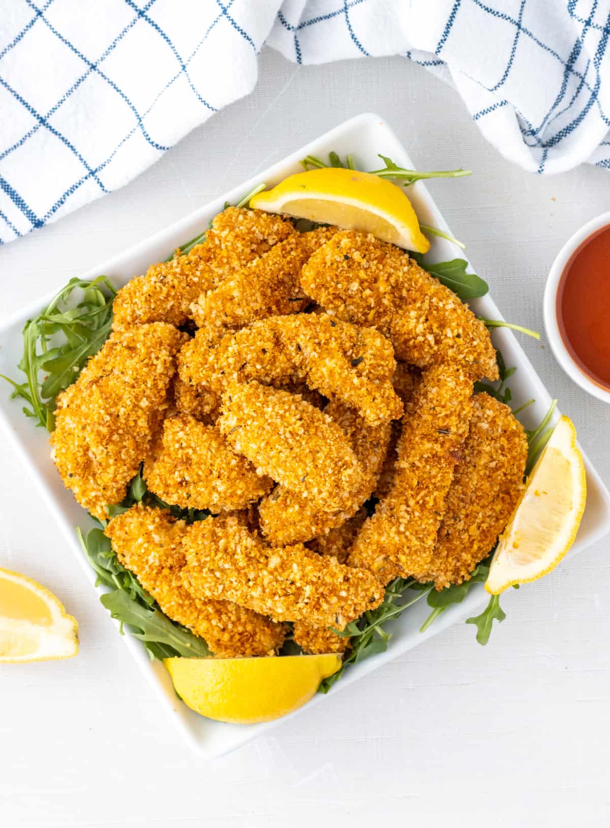 Panko crusted chicken strips on a plate with lemon wedges and a bowl of sauce on the side.