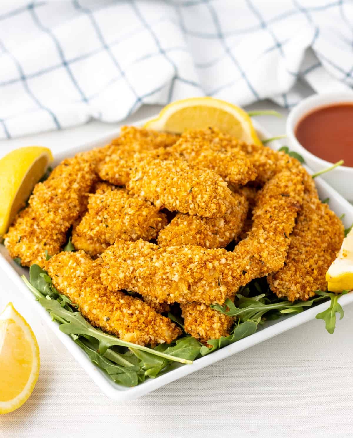 Panko crusted chicken strips on a plate with lemon wedges next to a bowl of sauce.