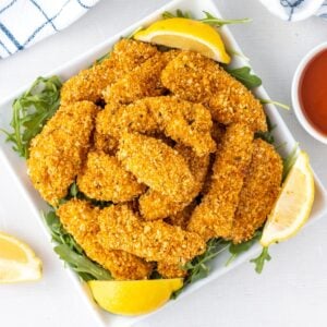 Panko crusted chicken on a plate with lemon wedges and a bowl of sauce to the side.