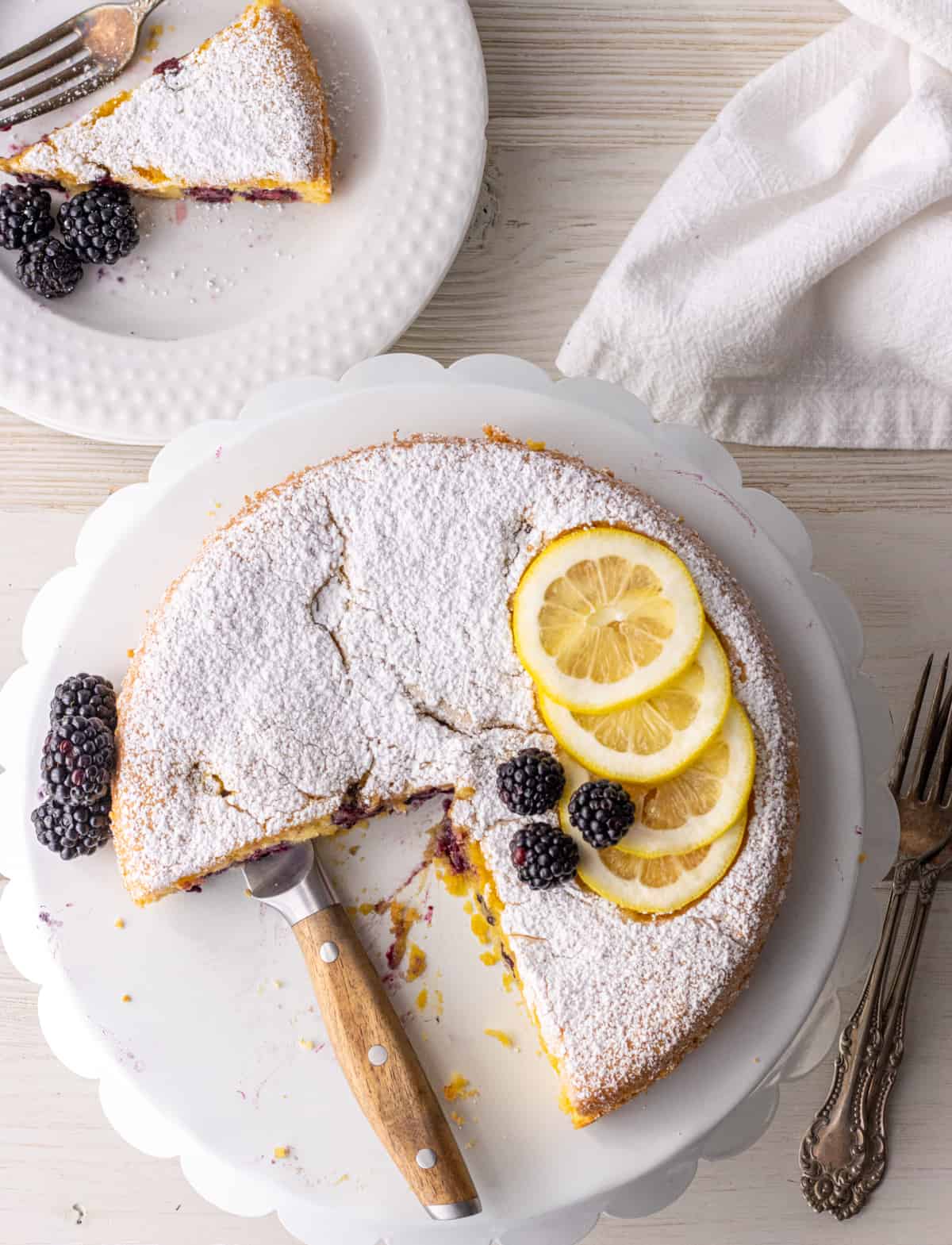 Blackberry almond cake on a cake stand with a knife and a slice on a place to the side.