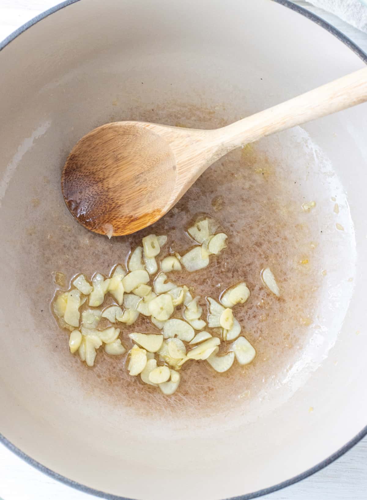 Sliced garlic simmering in oil in a pot with a wooden spoon.
