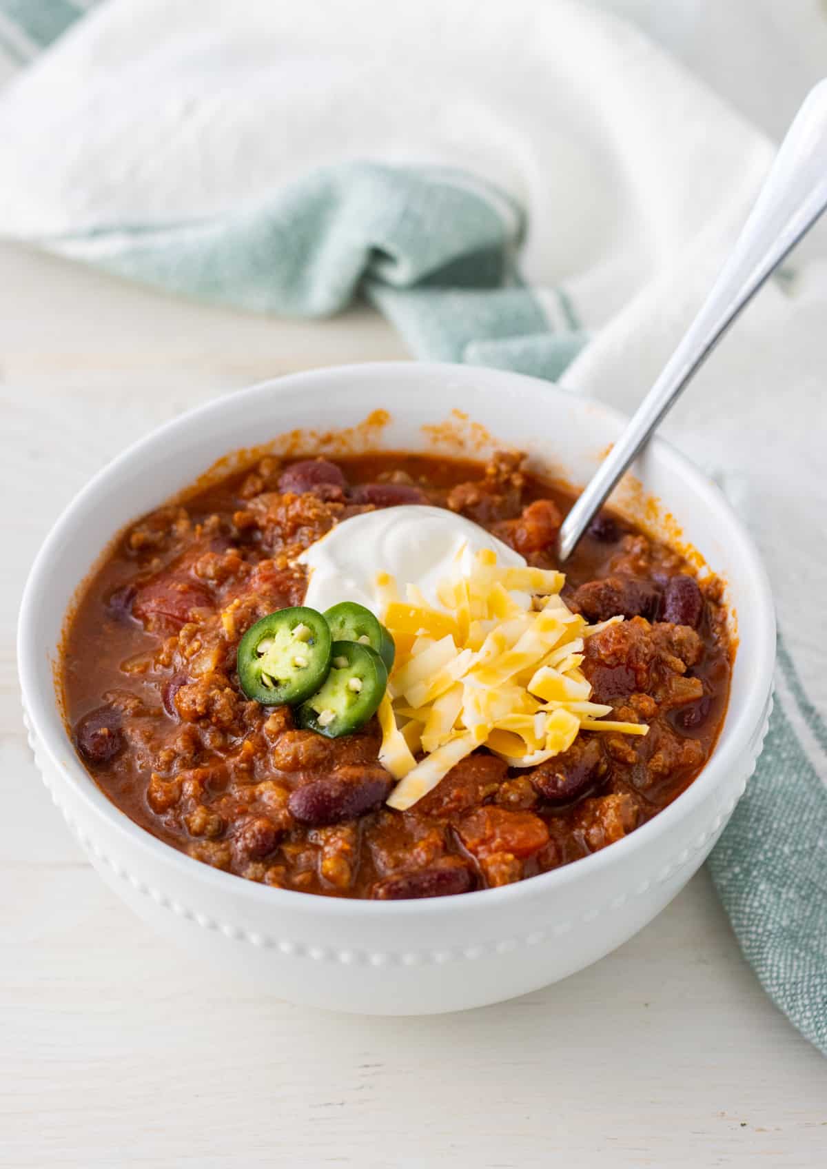 Bowl of chili with a spoon topped with sour cream, shredded cheese, and sliced jalapeños.