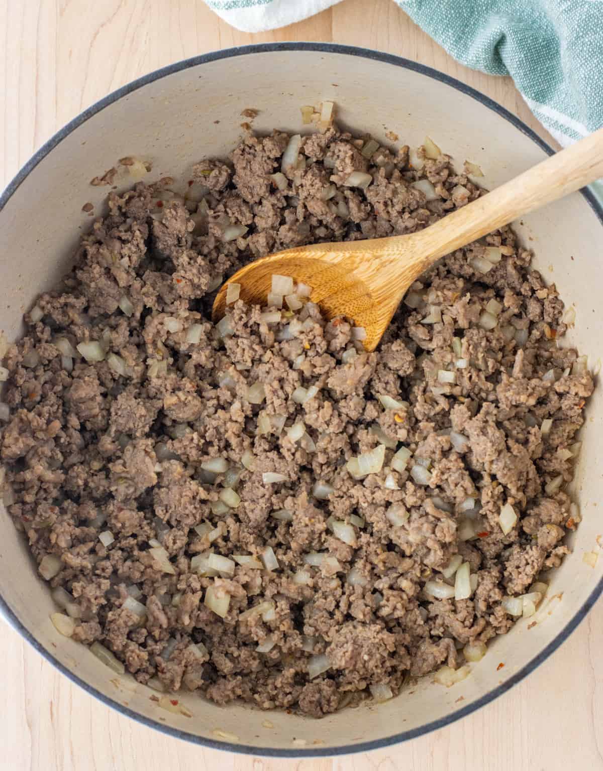 Cooked ground beef, sausage, and diced onions in a Dutch oven with a wooden spoon.