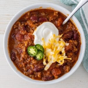 Bowl of chili with a spoon topped with sour cream, shredded cheese, and jalapeno slices.