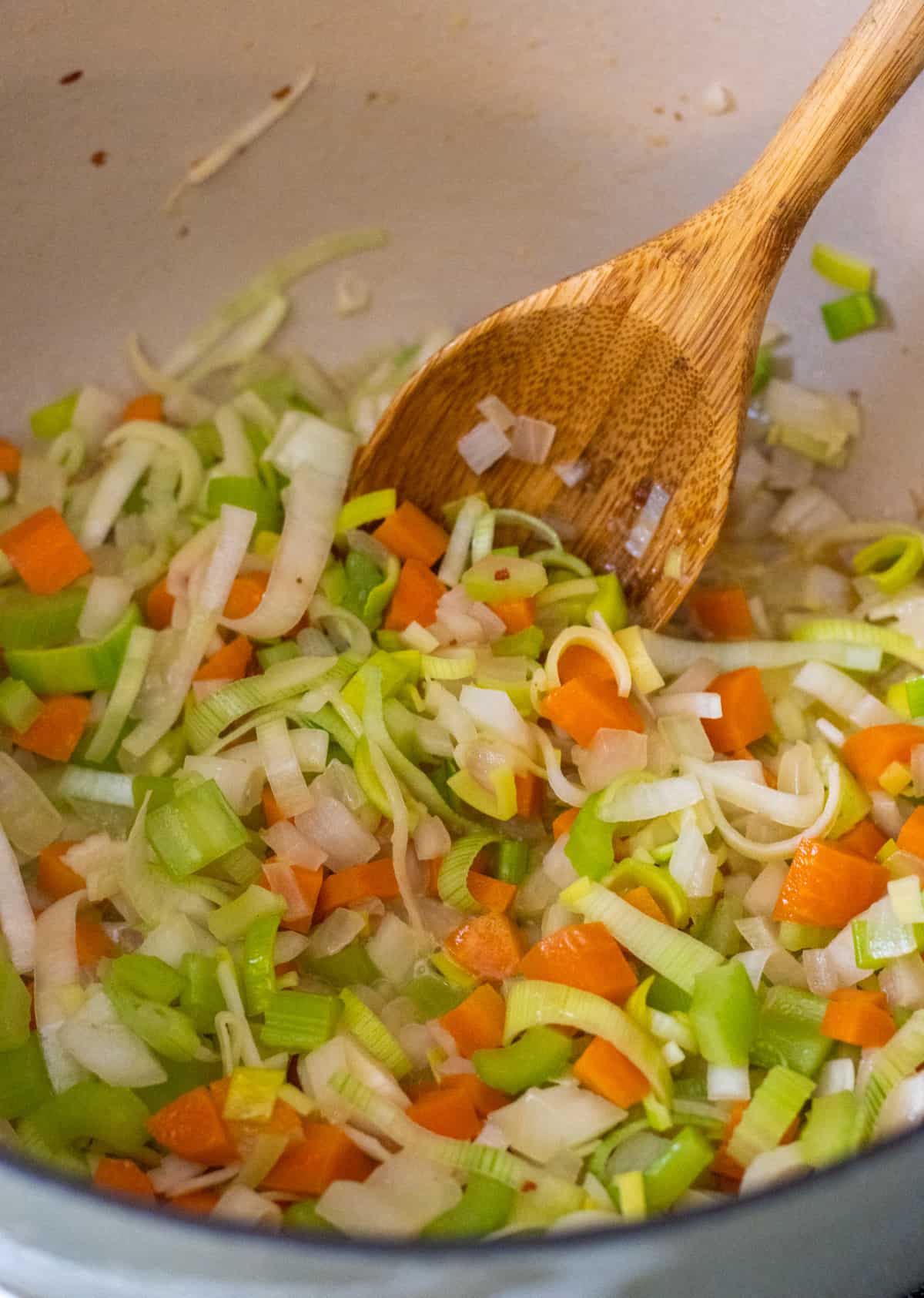 Sliced carrots, celery, onion, leeks, and garlic in a pot with a wooden spoon.
