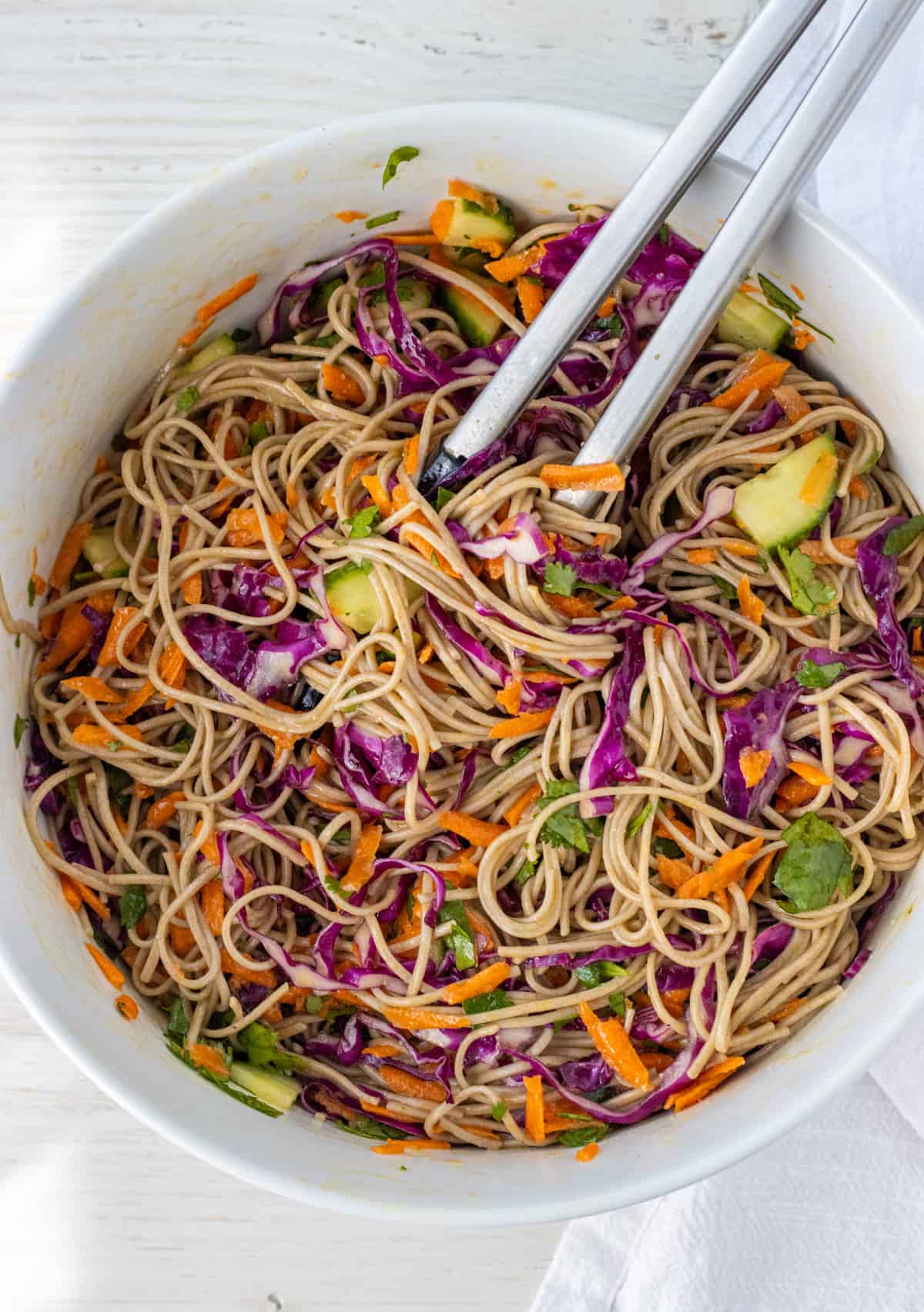 Soba noodle salad tossed in miso dressing in a large bowl with a pair of tongs.
