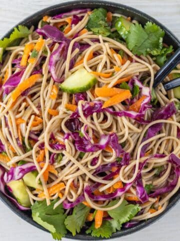 Bowl of soba noodles with sliced red cabbage, carrots, cucumbers, and chopped parsley with two chopsticks.