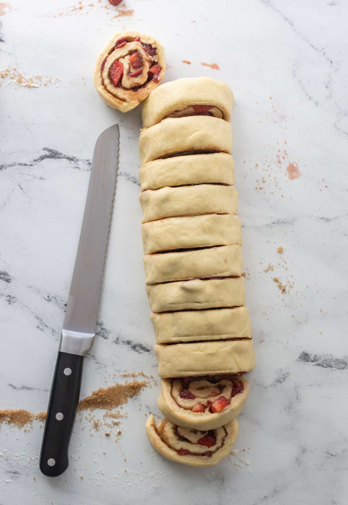 Strawberry cinnamon roll log cut into slices next to a serrated knife.