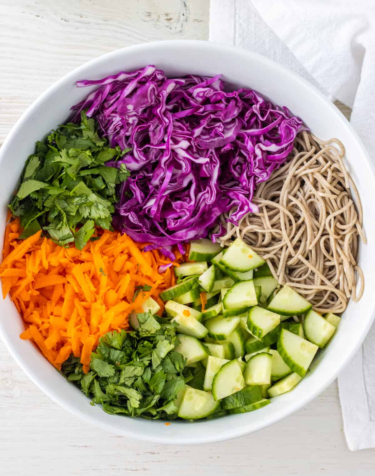 Sliced purple cabbage, sliced cucumbers, shredded carrots, and chopped parsley and cilantro in a large bowl with soba noodles.