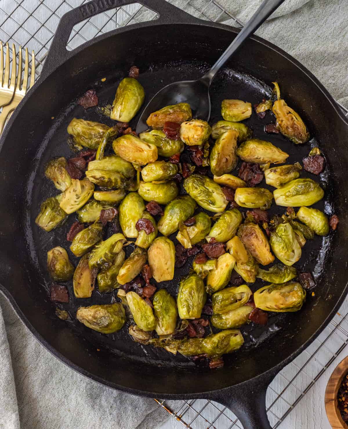 Caramelized Brussels sprouts and bacon in a cast iron skillet with a large spoon on top of a wire rack and towel with forks on the side.