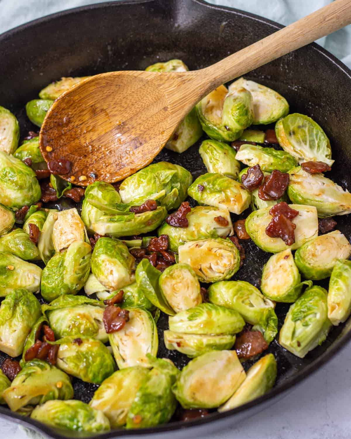 Halved Brussels sprouts and bacon tossed in a brown sugar sauce in a cast iron skillet with a wooden spoon.
