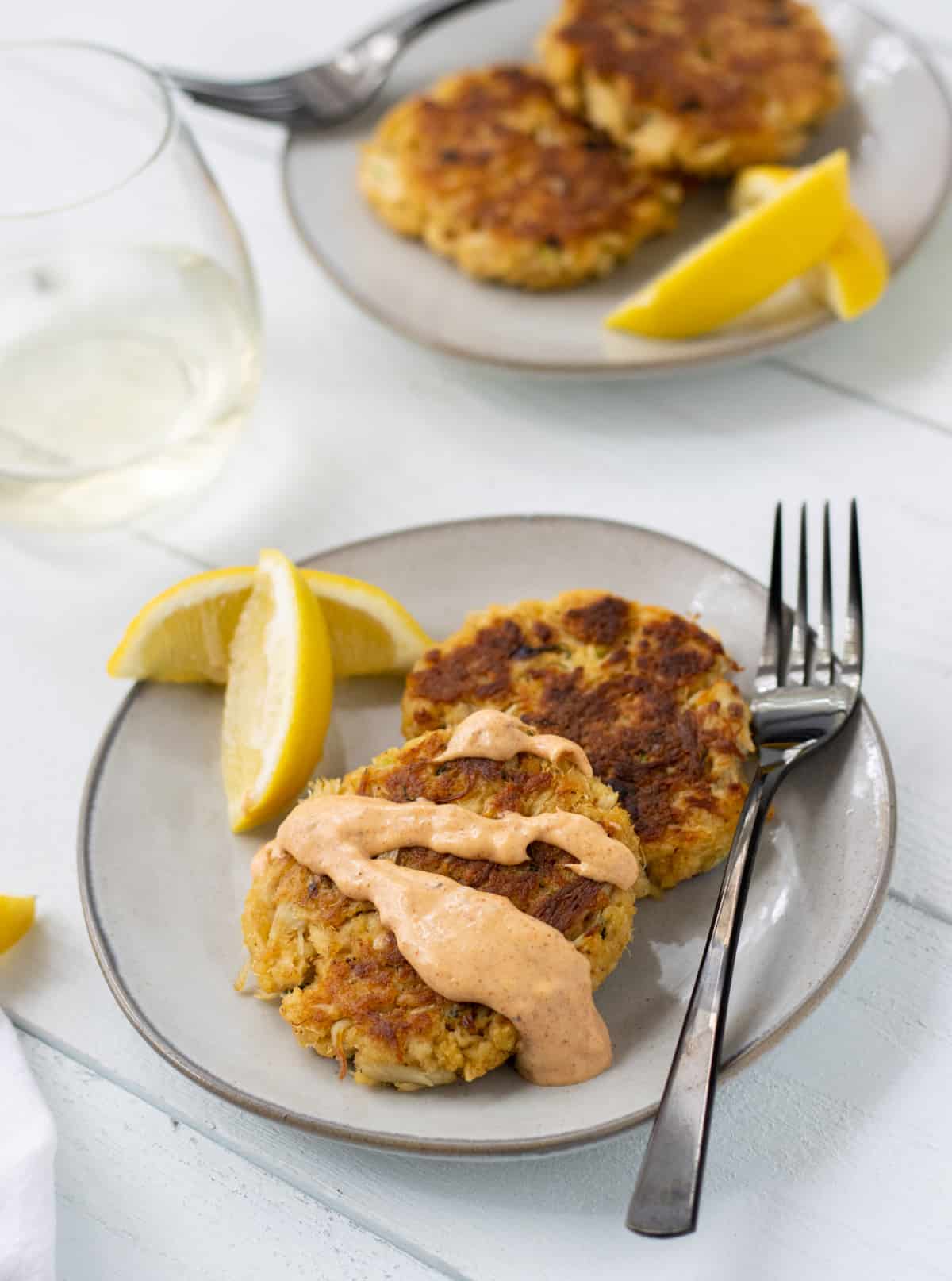 Two crab cakes on a plate with remoulade sauce drizzled on one with a fork and lemon wedges.