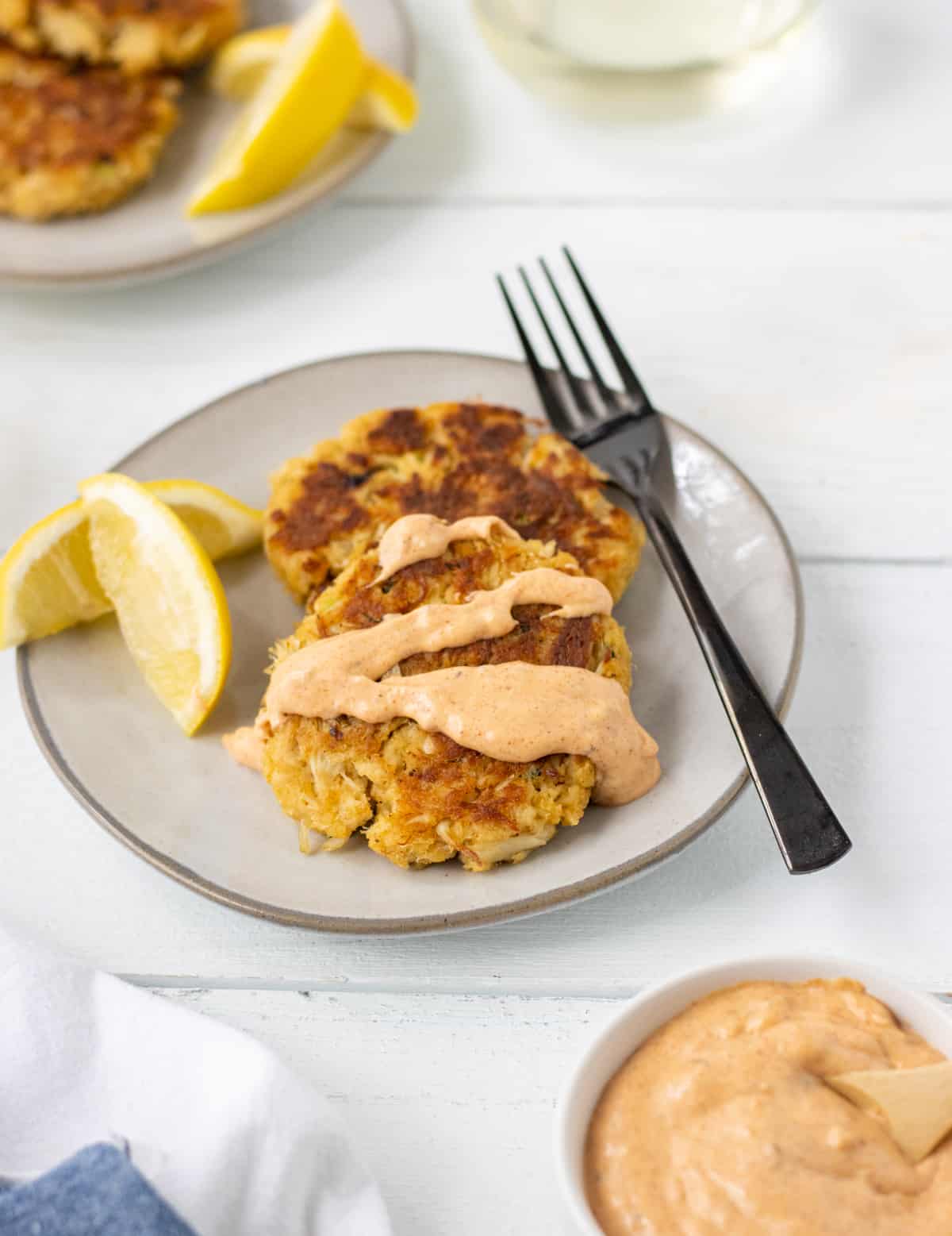 Two crab cakes topped with remoulade sauce on a plate with a fork and lemon wedges and another plate in the background.