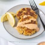 Crab cakes on a plate with lemon wedges and a fork and topped with remoulade sauce.