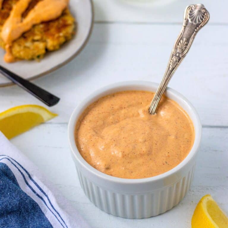 Remoulade sauce in a bowl with a spoon and crab cakes and lemon wedges on the side.