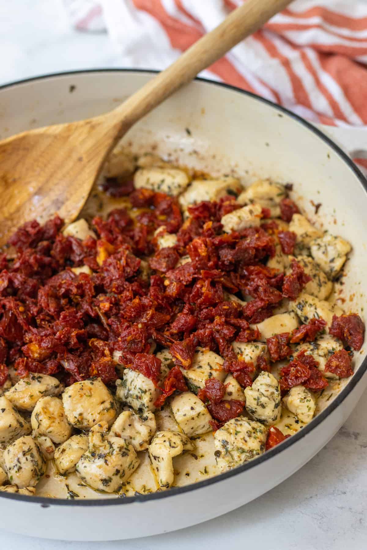 Sun-dried tomatoes in a skillet with cooked chicken and a wooden spoon.