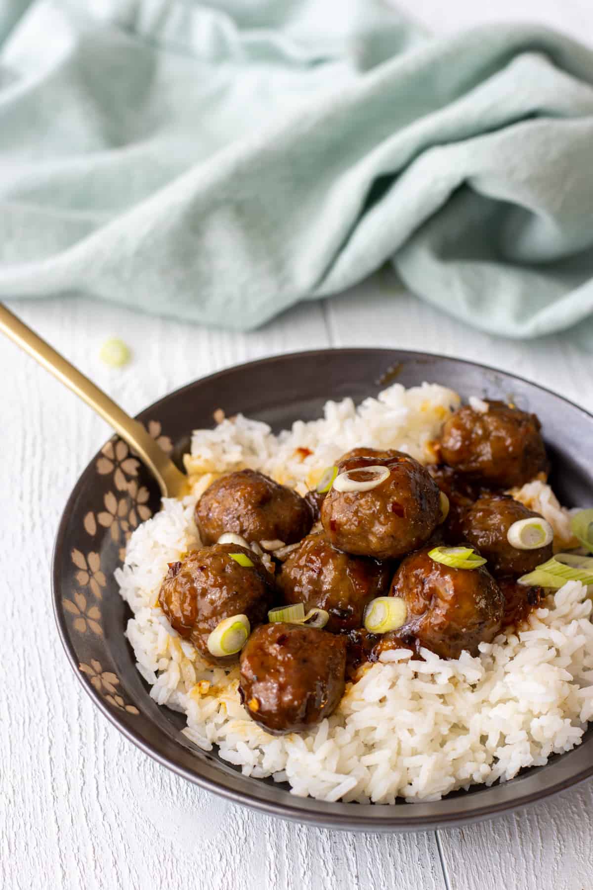 Sweet and spicy meatballs on white rice topped with sliced green onions with a fork and a towel.
