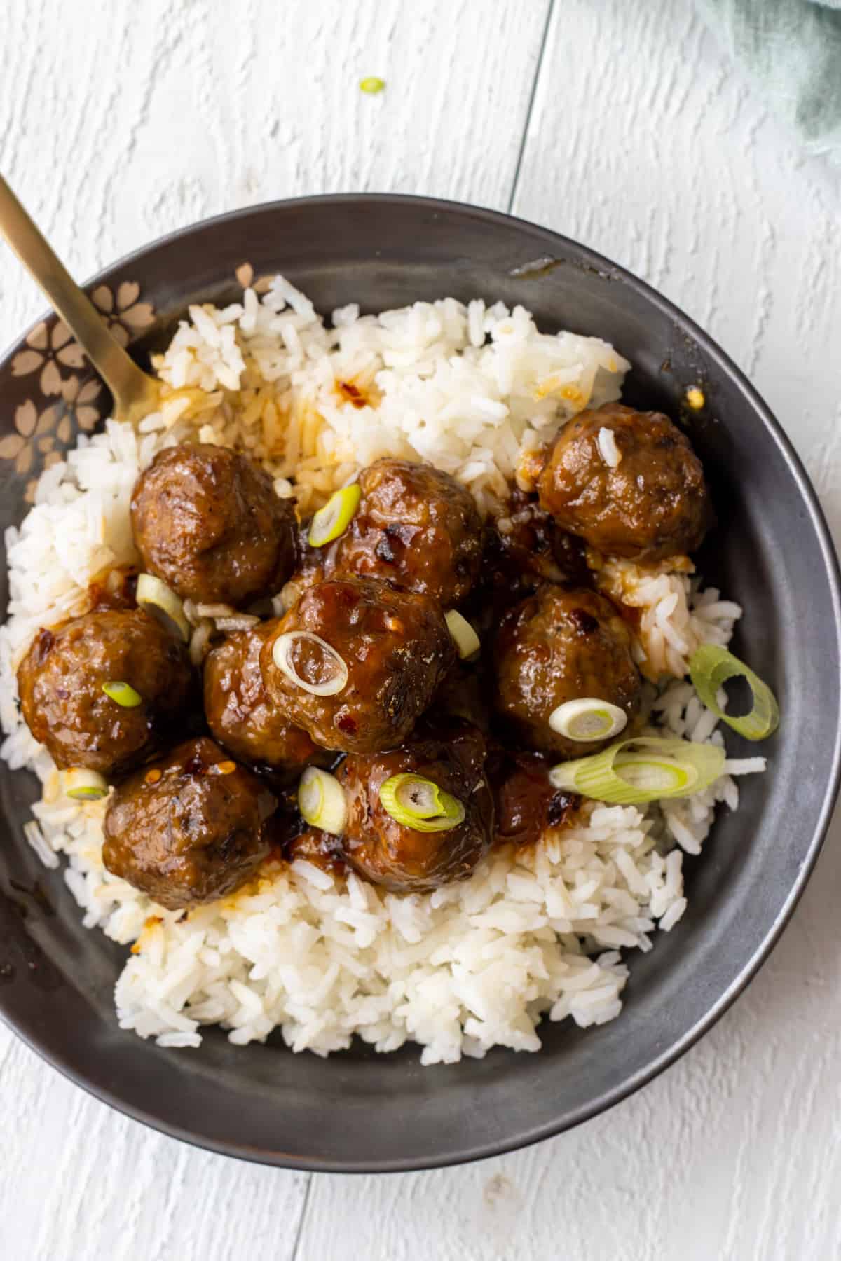 Sweet and spicy meatballs on top of white rice with sliced green onions, a fork, and a towel on the side.