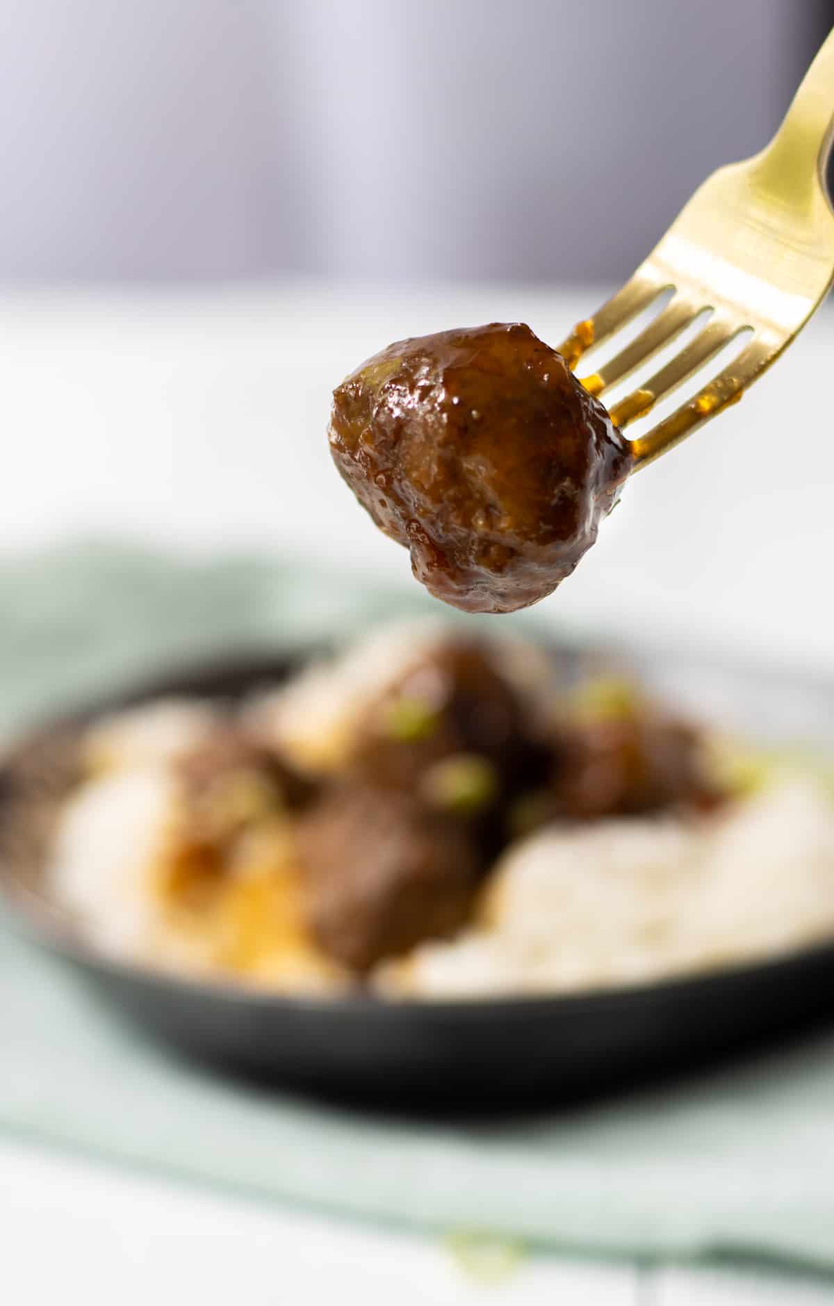A meatball on a fork held in the air with a plate of rice and meatballs in the background.