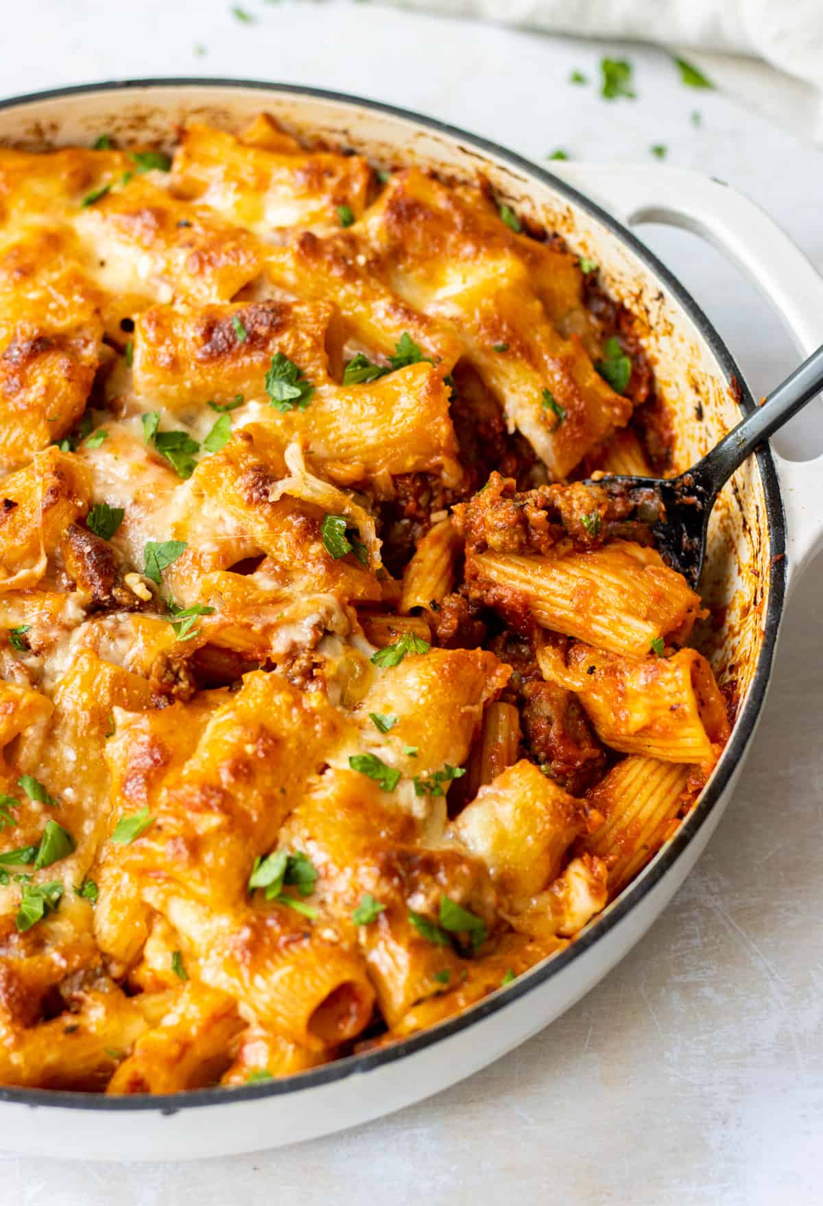 Baked rigatoni with sausage in a skillet with a scoop missing and a large spoon in the spot.