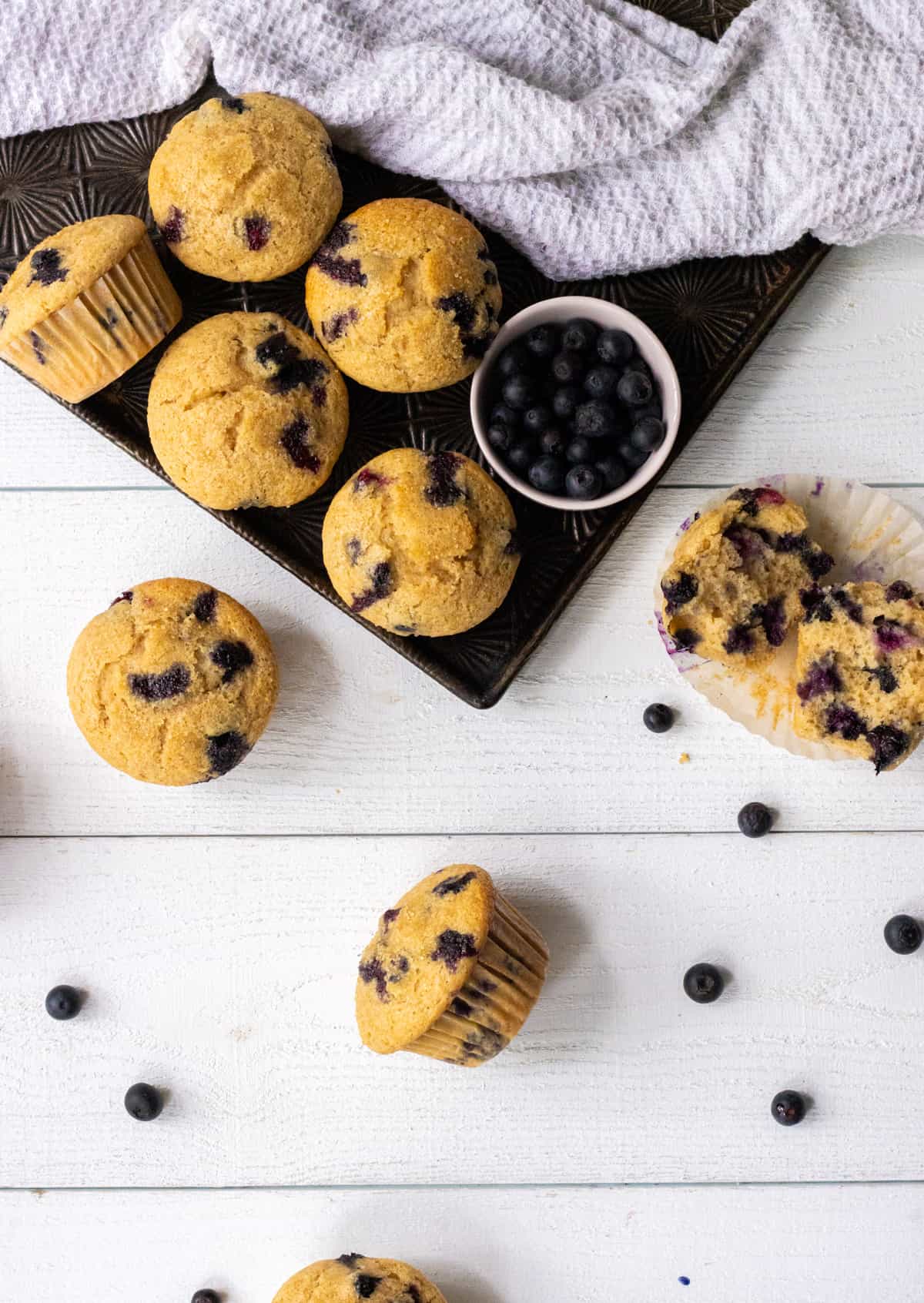 Blueberry buttermilk muffins on a cookie sheet with a bowl of blueberries and a tea towel and more muffins and blueberries on the table
