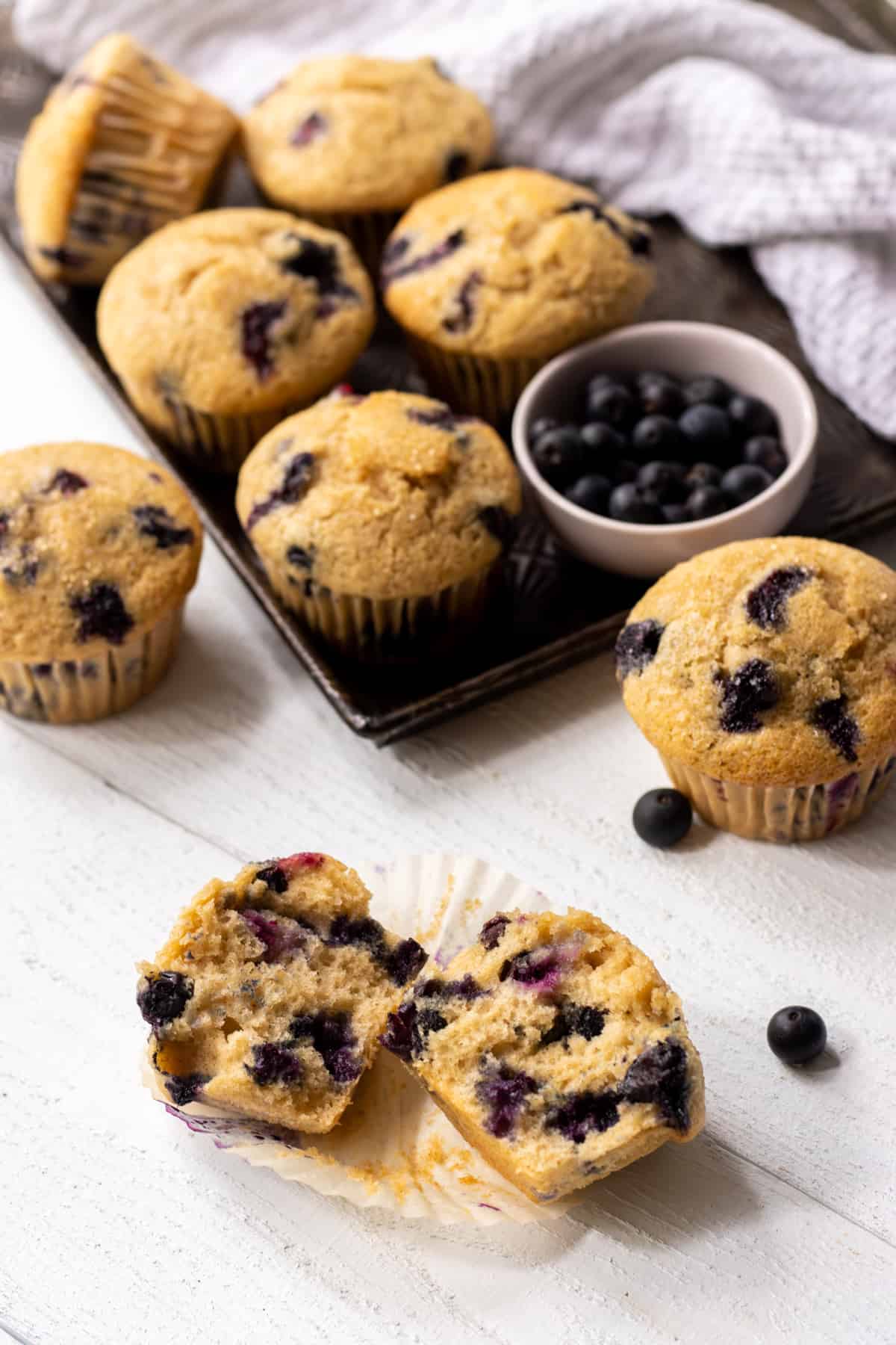 Blueberry buttermilk muffins on a cookie sheet with a bowl of fresh blueberries and more muffins on the table with one in half.
