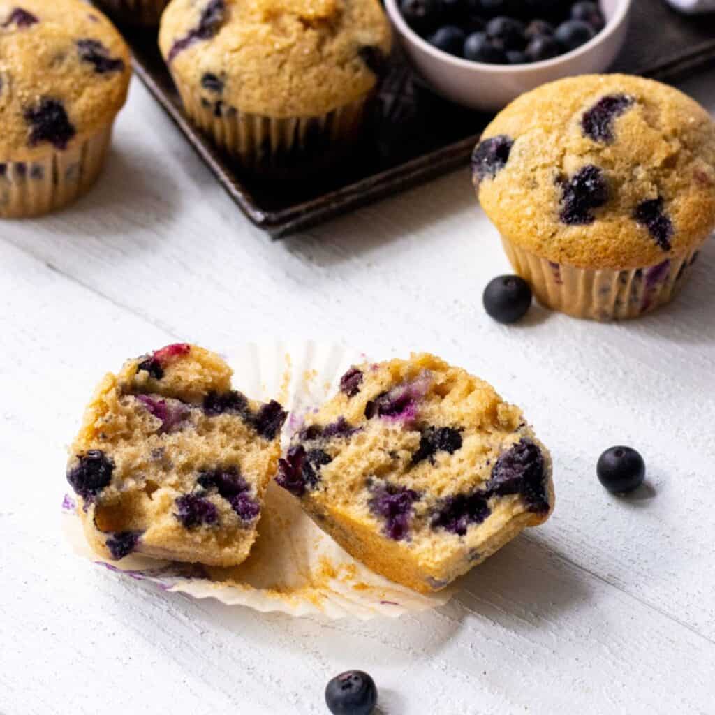 Blueberry buttermilk muffins with one split in half and more on a baking sheet in the back with blueberries all around.