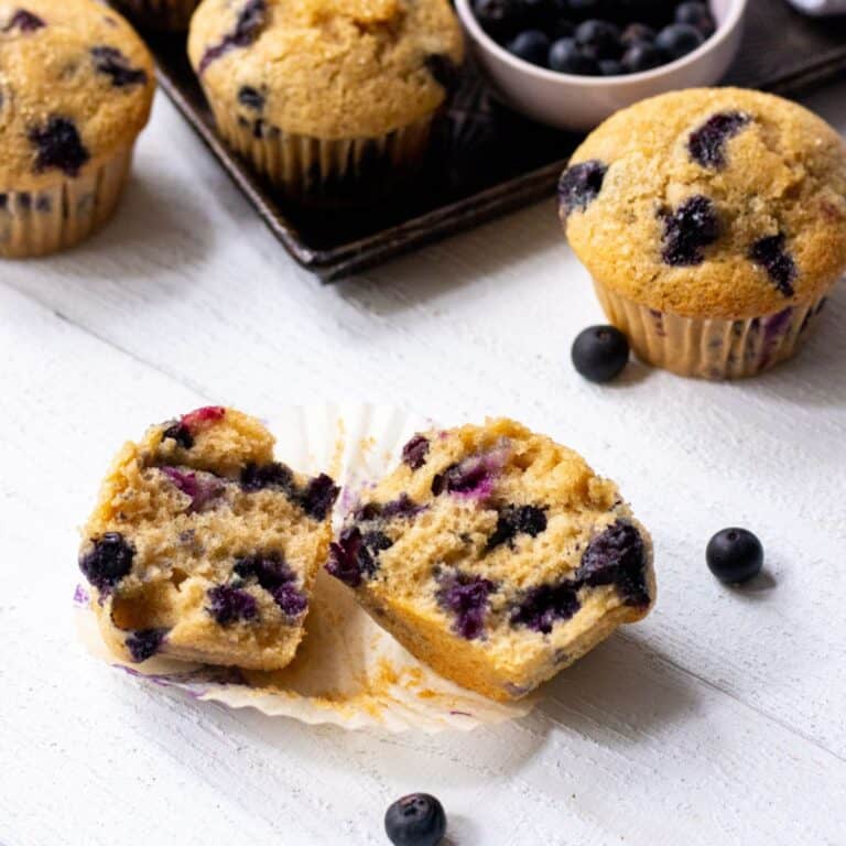 Blueberry buttermilk muffins with one split in half and more on a baking sheet in the back with blueberries all around.