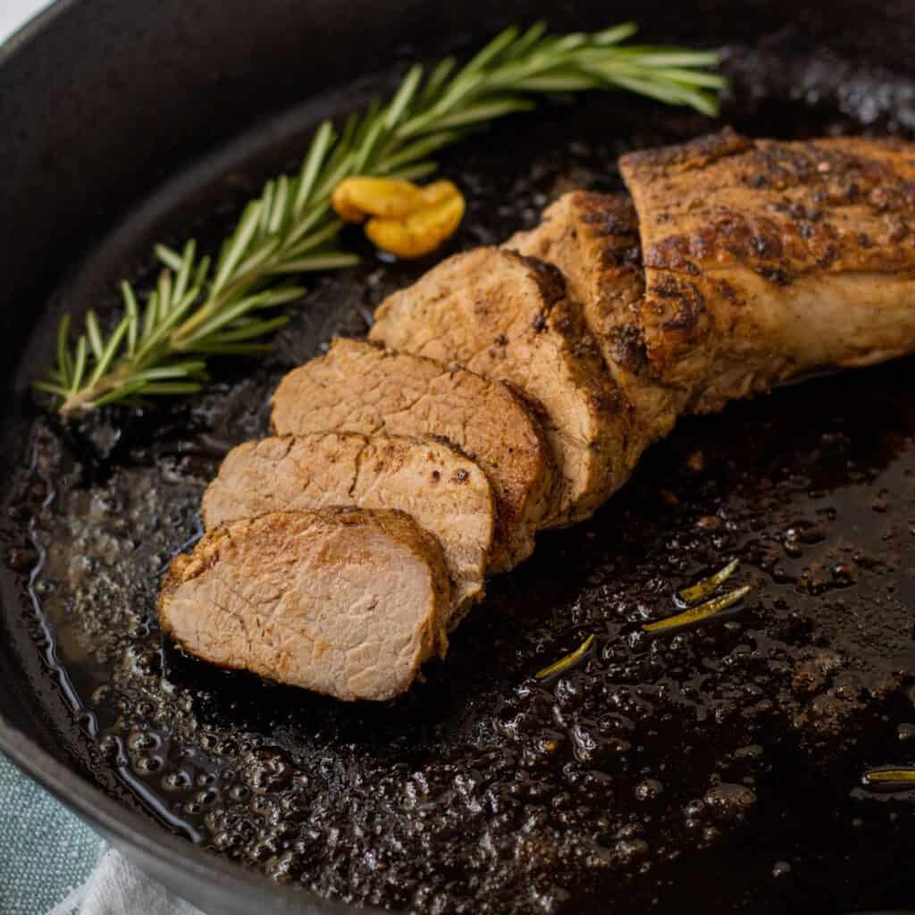 Sliced pork tenderloin in a cast iron skillet with a rosemary sprig and a caramelized garlic clove.