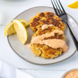 Two crab cakes with remoulade sauce on a plate with lemon wedges and a fork.