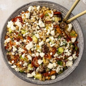 Mediterranean farro salad topped with feta crumbles in a bowl with two gold spoons.