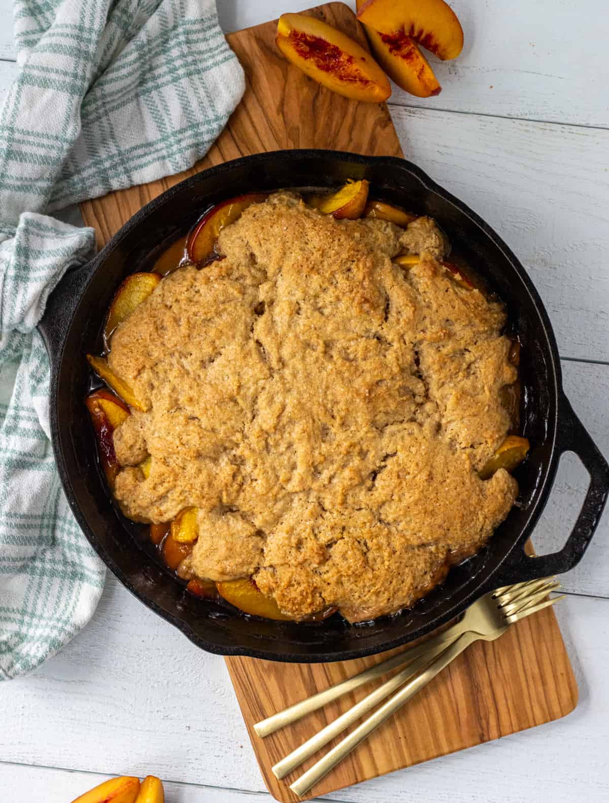 Peach cobbler in a cast iron skillet on a board with fresh peach slices and a pile of forks.