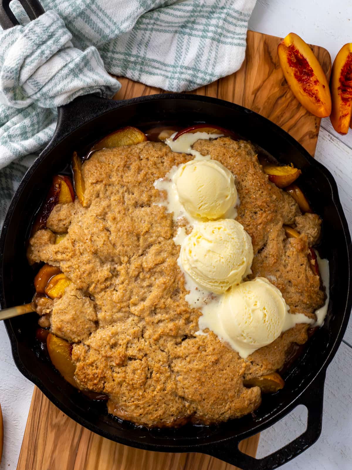 Peach cobbler in a cast iron pan topped with three scoops of vanilla ice cream on a wood board with fresh slices of peaches.