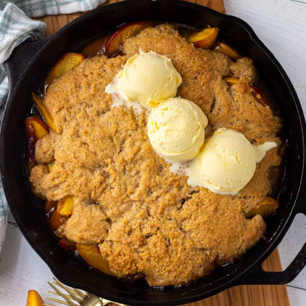 Peach cobbler in a cast iron skillet topped with three scoops of vanilla ice cream.