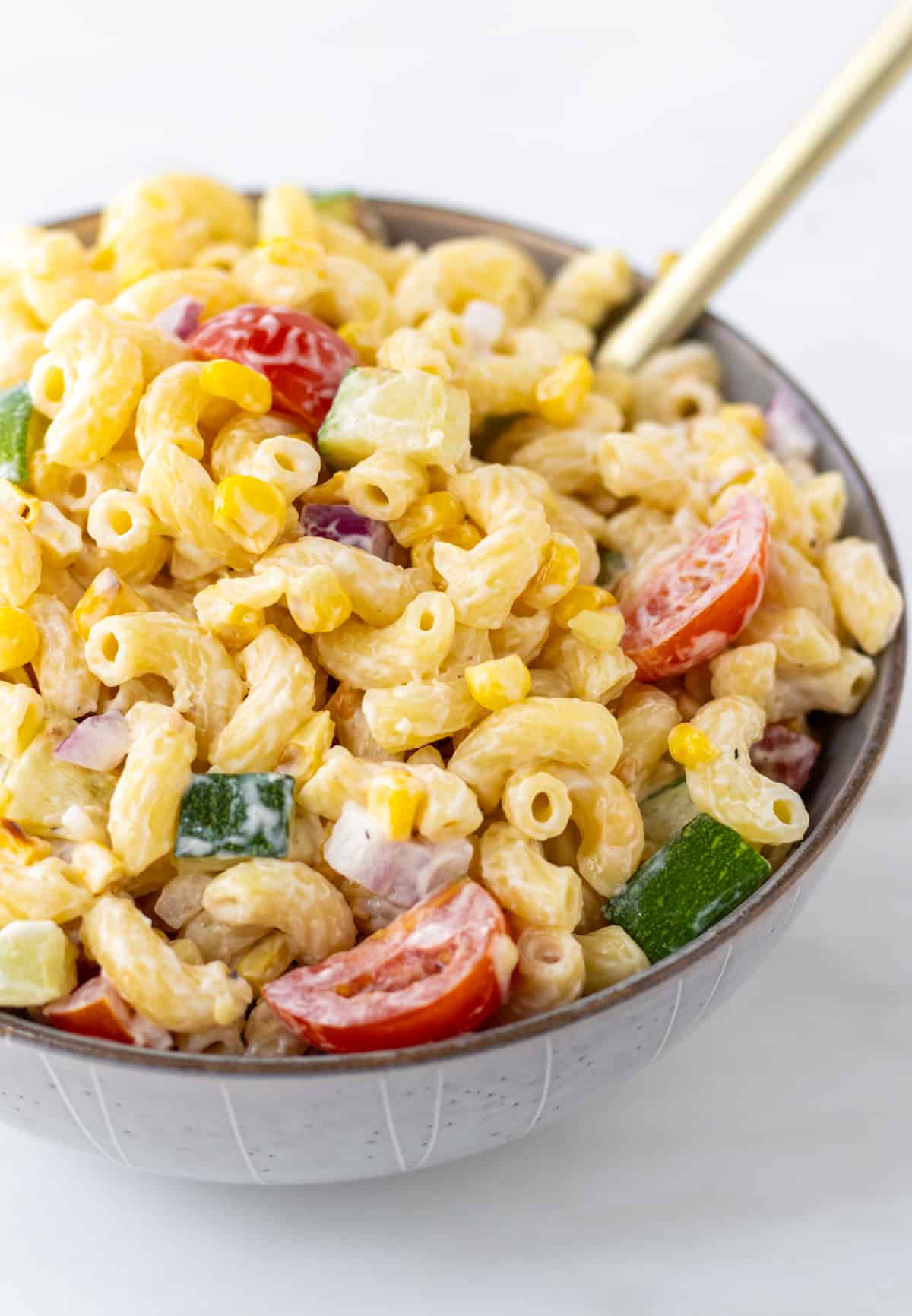 Creamy pasta salad with corn, zucchini, tomatoes, and red onion in a bowl with a spoon.