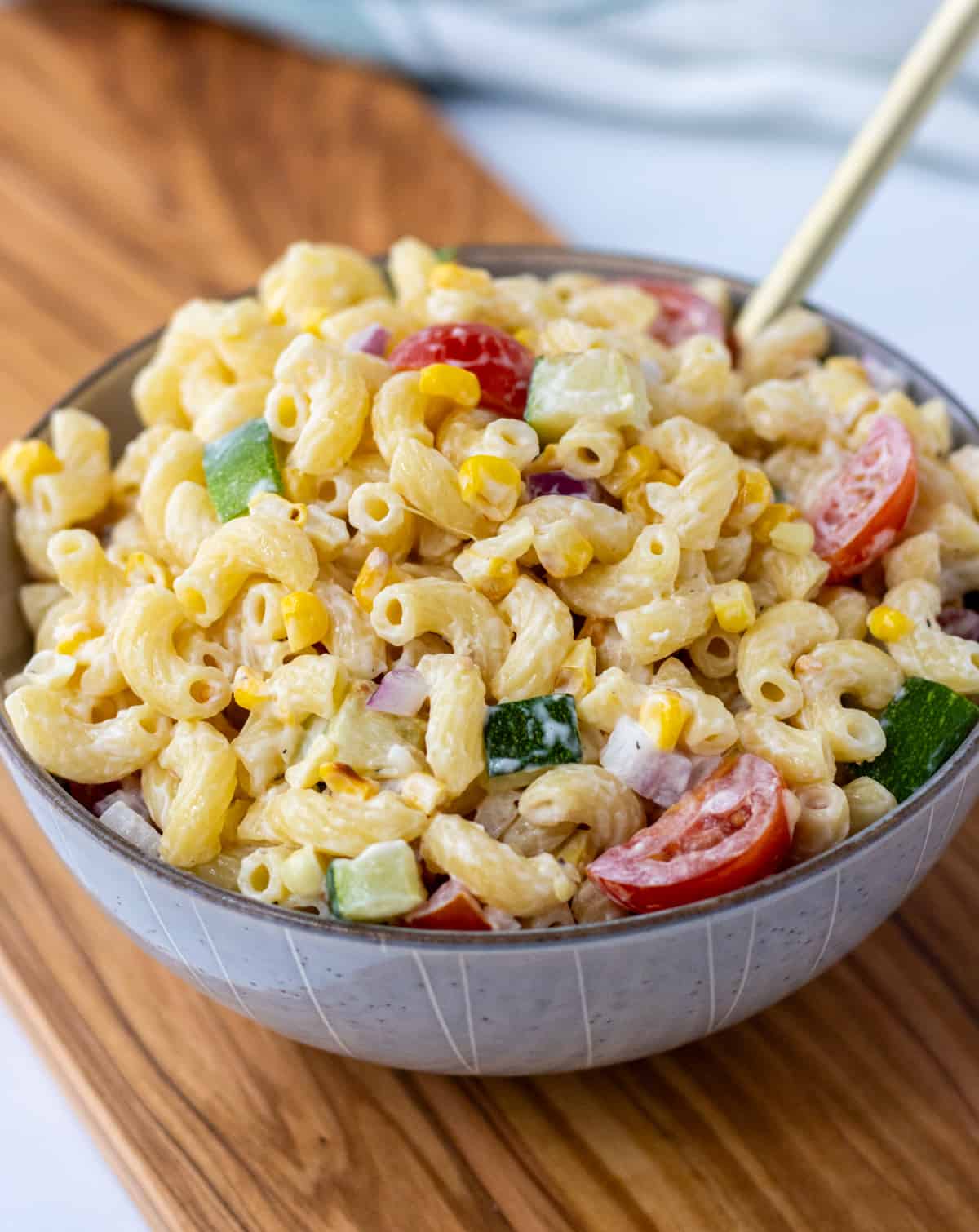 Creamy corn and zucchini pasta salad in a bowl with a spoon on top of a wood board.