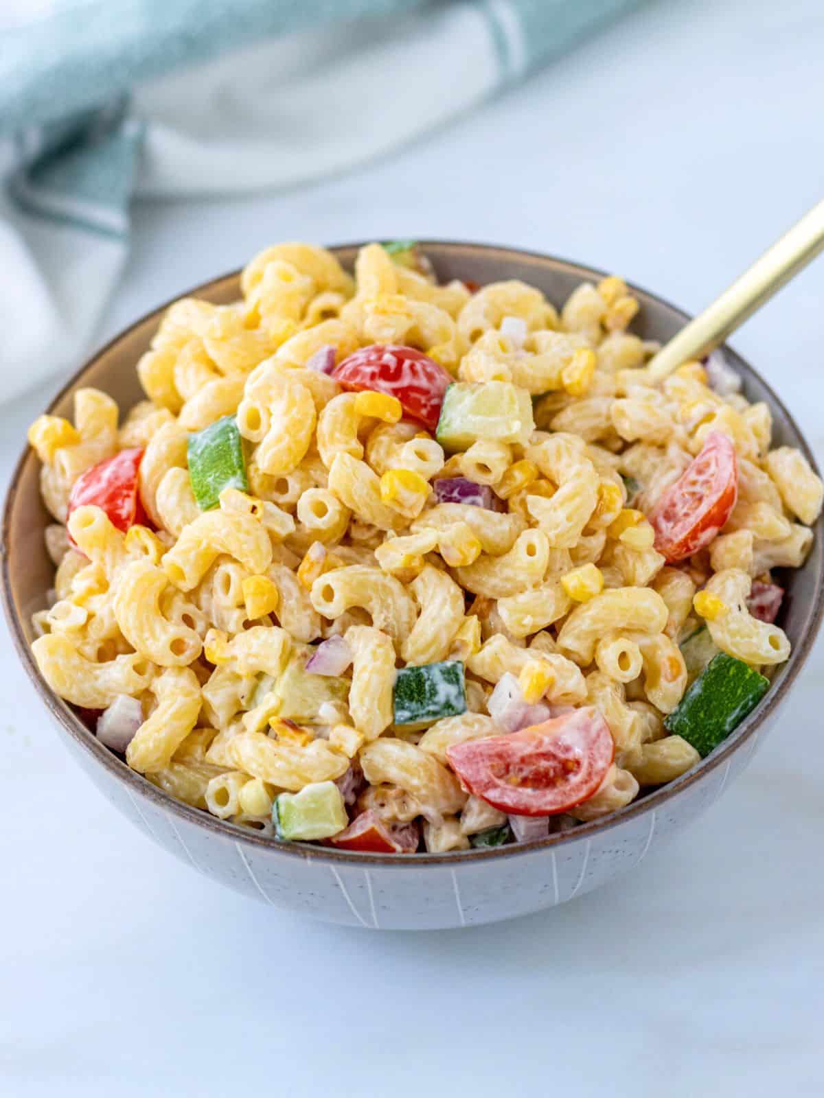 Creamy pasta salad with corn, zucchini, tomatoes, and red onion in a bowl.