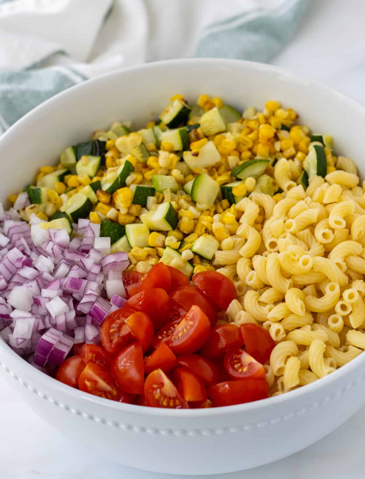 Quartered cherry tomatoes, diced red onion, charred corn and zucchini, and elbow noodles in a large bowl.