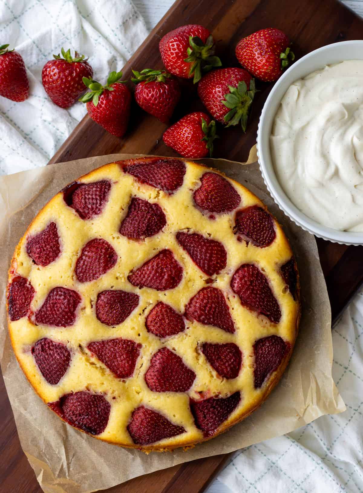 Strawberry studded one layer vanilla cake on a wood platter next to a bowl of whipped cream and fresh strawberries.