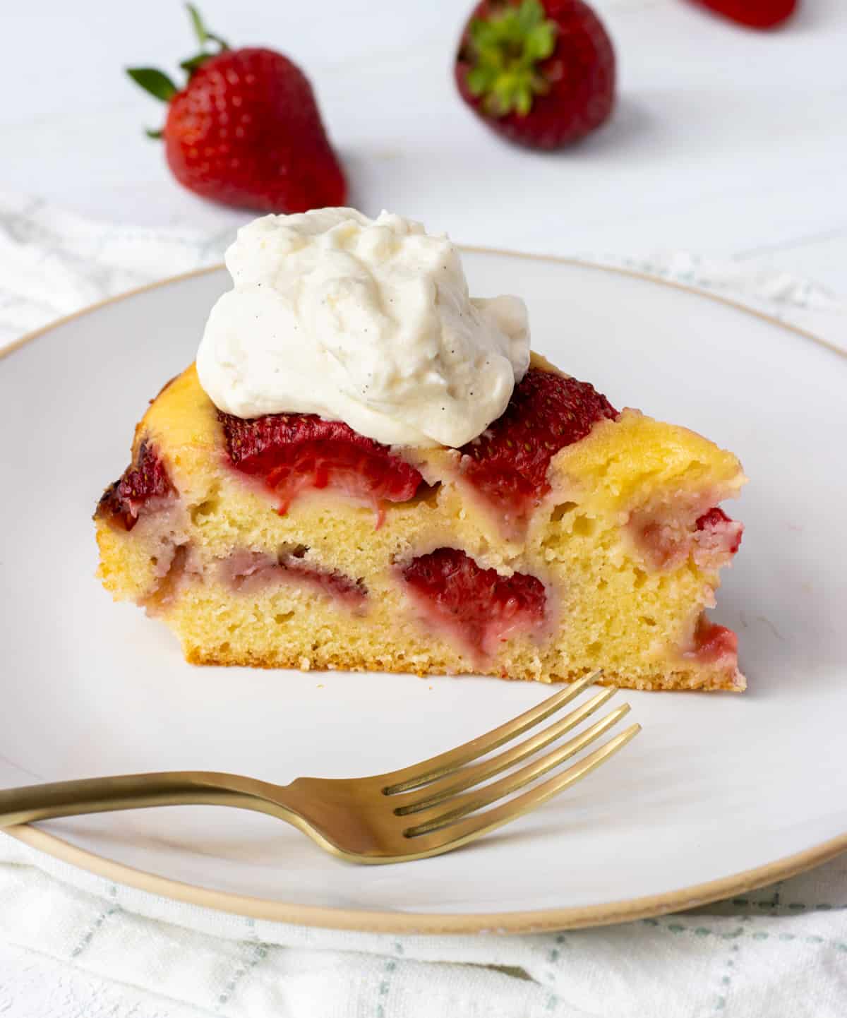 Slice of strawberry cake topped with whipped cream on a plate with a fork and fresh strawberries in the back.