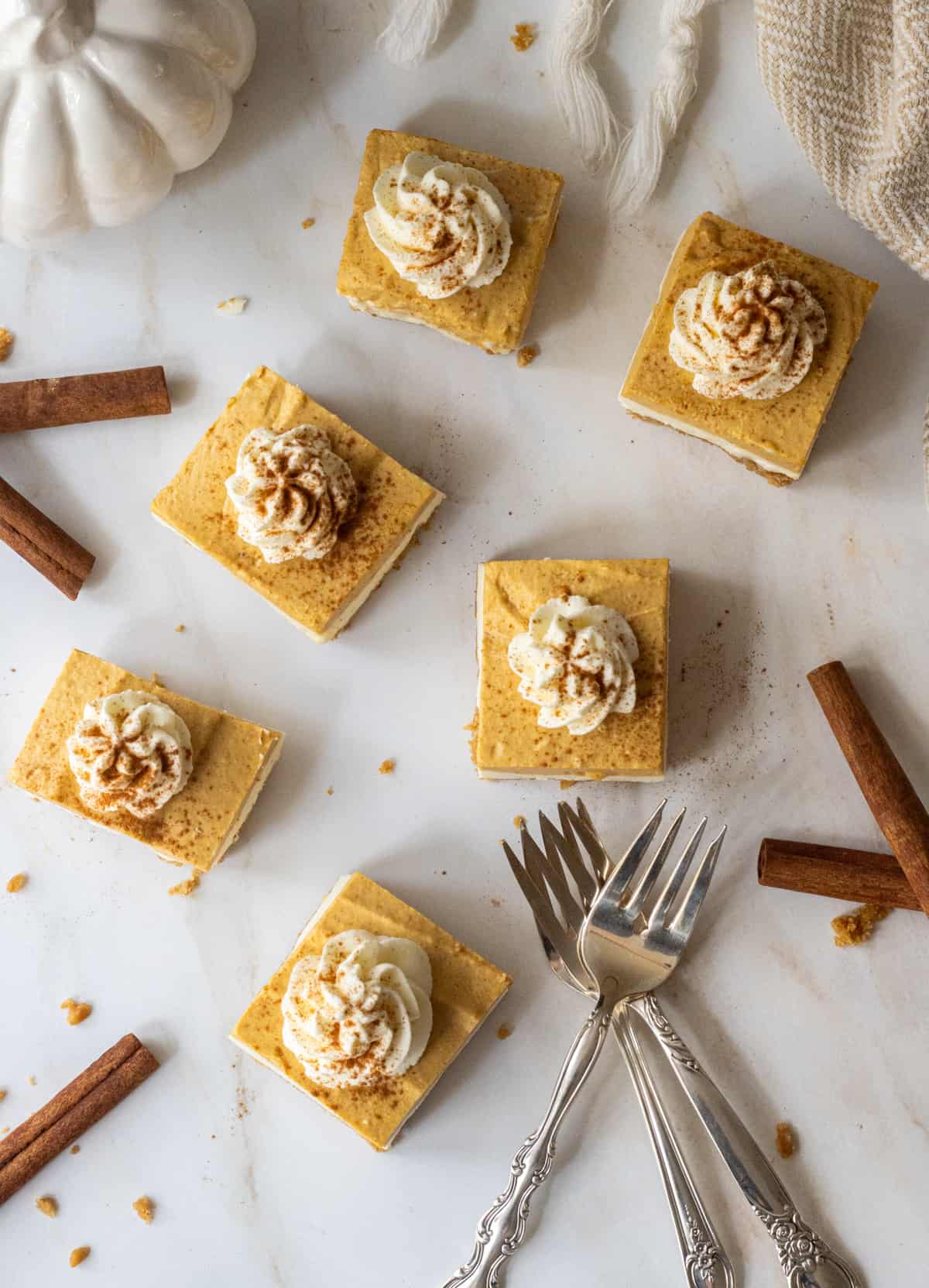Pumpkin cheesecake bars topped with whipped cream and cinnamon next to cinnamon sticks, forks, and a ceramic pumpkin.