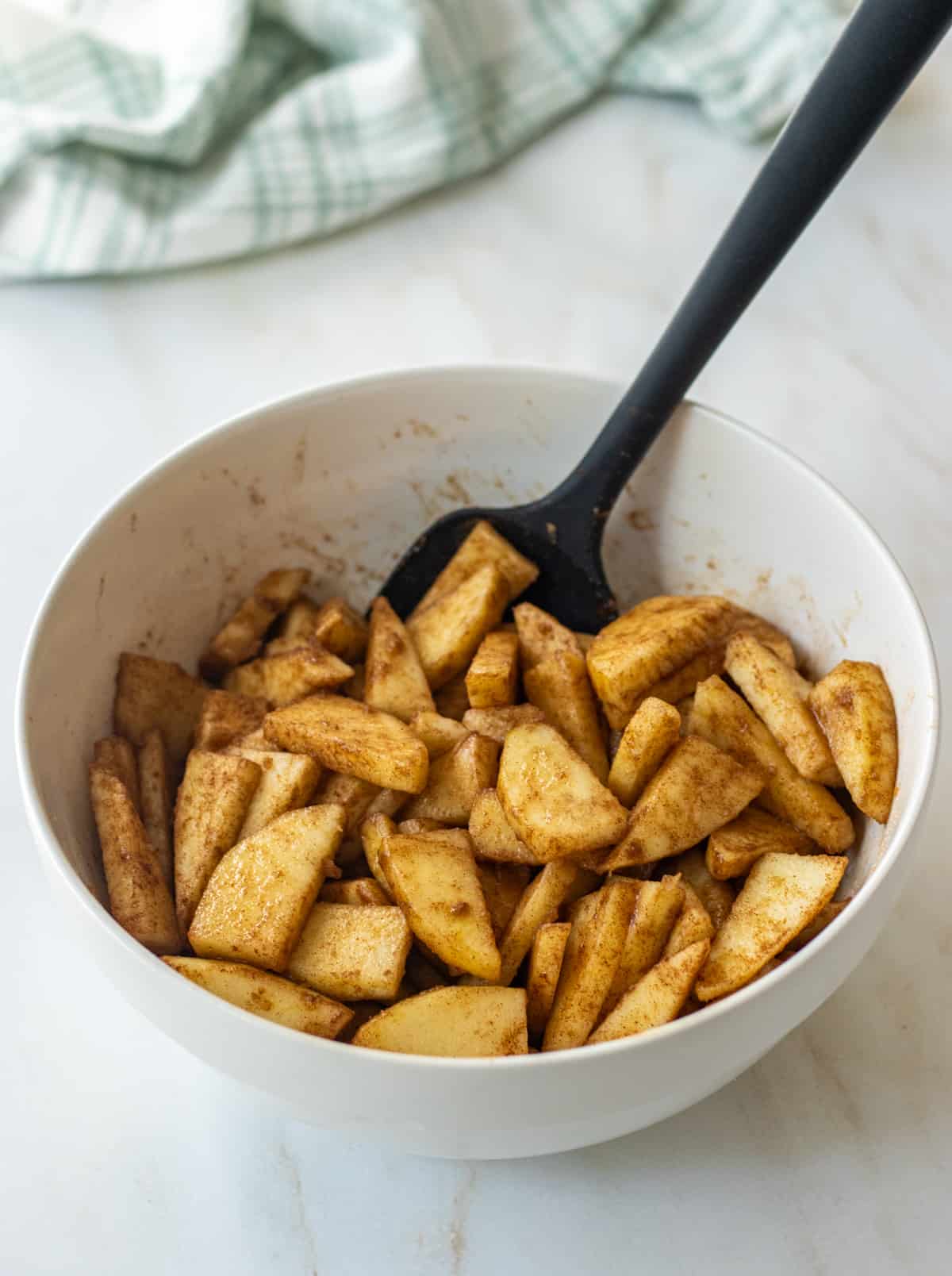Apple slices tossed with brown sugar and cinnamon in a bowl with a rubber spatula.