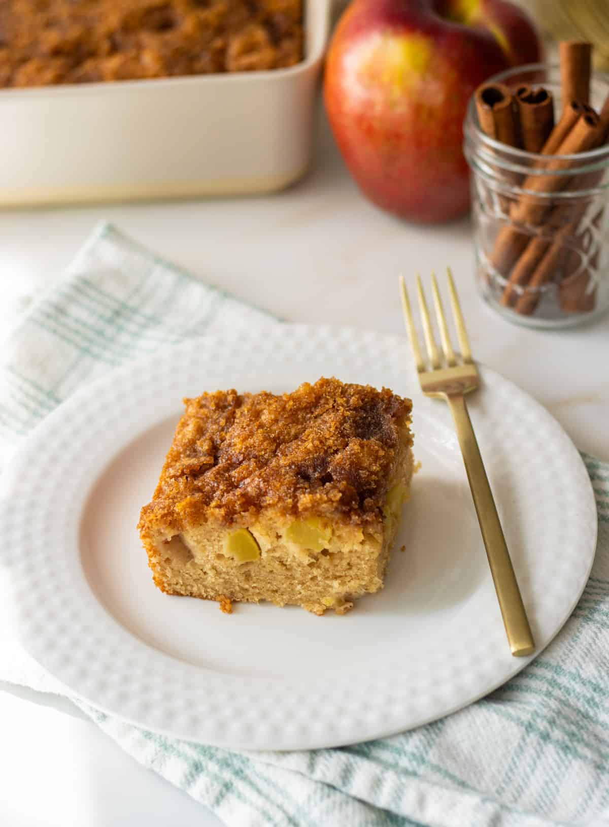 Slice of cinnamon apple cake on a plate with a fork on top of a towel.
