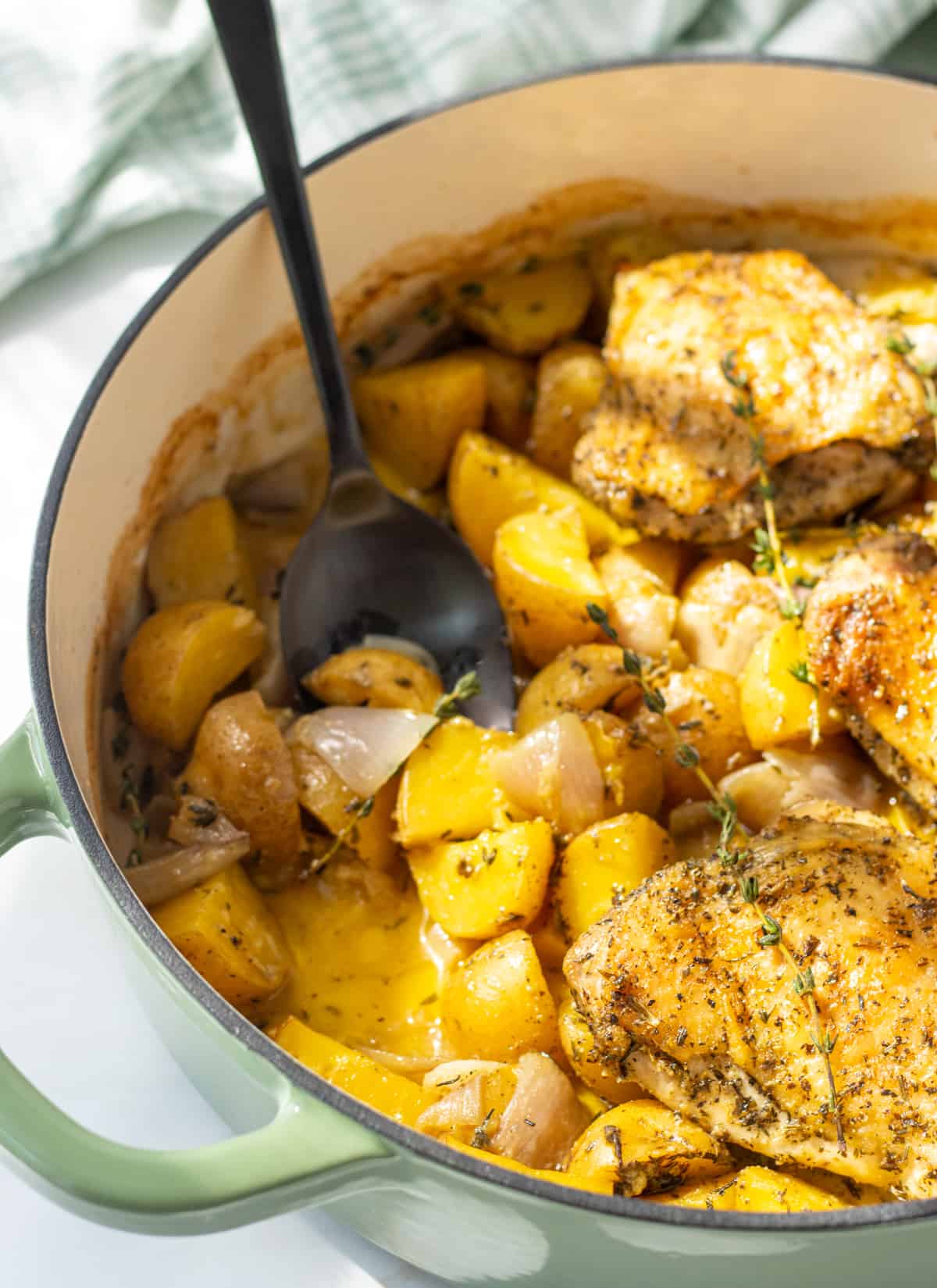 A large skillet with roasted potatoes and chicken thighs with a large spoon scooping a few potatoes.